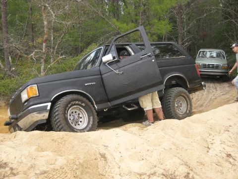 1994 Ford Bronco - A black 1994 Ford Bronco is stuck in the sand with its driver's door open, while another vehicle waits behind it.