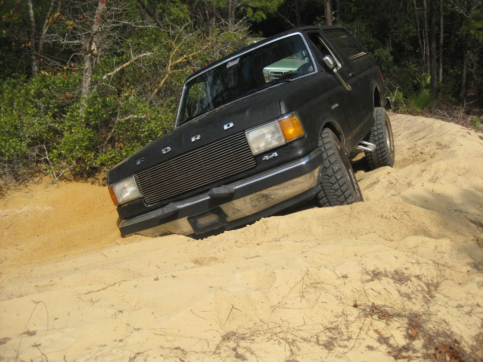 Black Ford Bronco with lifted suspension driving on a sandy hill. Grille has 'FORD' lettering