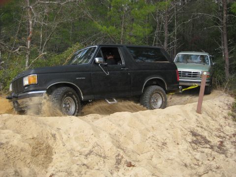 1994 Ford Bronco - A black Ford Bronco struggles in sandy terrain while being towed by a green Ford F-Series truck in a wooded area.