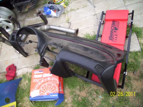 1995 Ford Probe - Various automotive parts, including a dashboard and exhaust components, are laid out on the ground next to a red creeper and a box labeled…