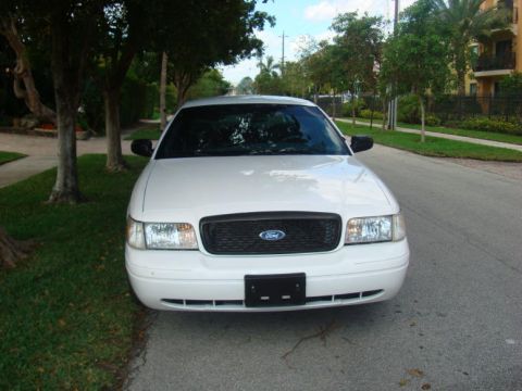 2002 Ford Crown Victoria - The front of a white 2002 Ford Crown Victoria is visible, parked on a street with trees and buildings in the background.