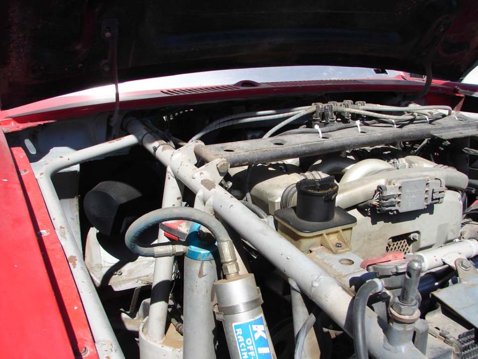 Red vehicle engine bay with roll cage, shock absorber, and various hoses and components visible