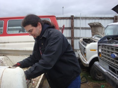 1989 Ford Bronco - A man works on the bed of a white truck, with other vehicles visible in the background under an overcast sky.