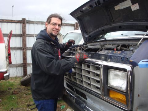 1989 Ford Bronco - Man works on the front grille of a blue Ford truck with the hood open. Another vehicle is visible to the left.