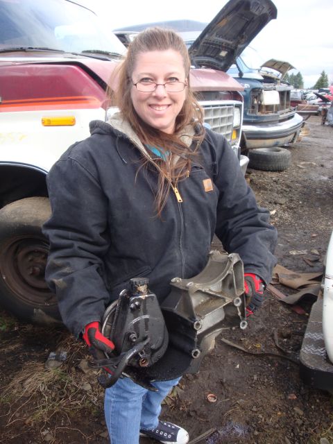 1989 Ford Bronco - A woman holds automotive parts in front of several vehicles at a junkyard.
