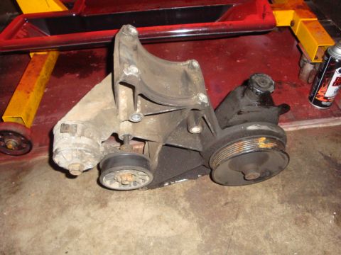 1989 Ford Bronco - A disassembled power steering pump assembly, including pulleys and mounting bracket, sits on a concrete floor.