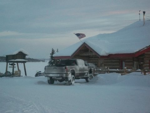 1986 Chevrolet Blazer - A silver pickup truck is parked in the snow outside a log cabin, with a frozen lake in the background.