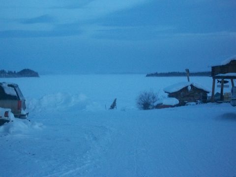 1986 Chevrolet Blazer - A tan SUV sits partially buried in snow beside a frozen lake, with a small cabin in the background.