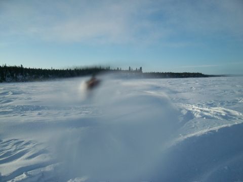 1986 Chevrolet Blazer - A snowmobile travels across a vast, snow-covered area, kicking up a cloud of snow against a backdrop of trees and a clear blue sky.