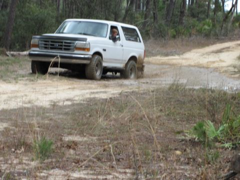 1990 Ford Bronco - A white Ford Bronco navigates a muddy trail surrounded by trees.