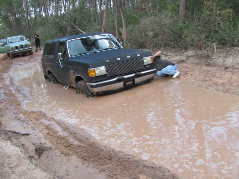 1990 Ford Bronco - A black Ford Bronco is stuck in a muddy puddle on a dirt road, with another vehicle visible in the background.