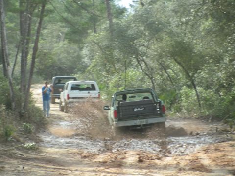 1990 Ford Bronco - Multiple trucks navigate a muddy path through a wooded area.