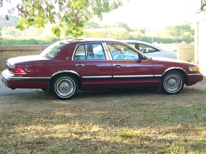 Side view of a dark red four-door sedan with chrome trim and lace-style wheels parked on gravel under trees.
