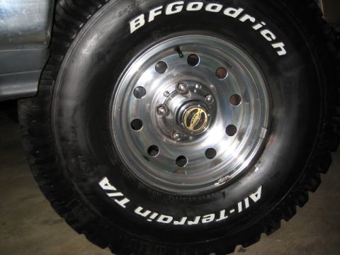 1990 Ford Bronco - Close-up of a polished aluminum five-lug wheel with ten round vent holes and a small gold-face center cap, mounted on a raised-white-letter…