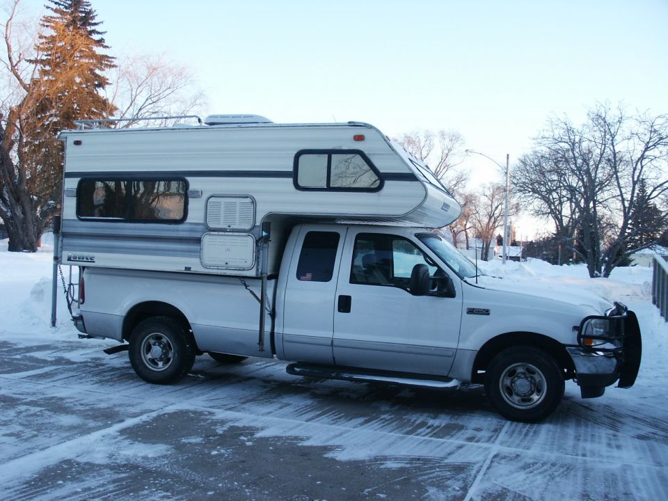 White pickup truck with a camper shell, parked in snow. Front grille and headlights are visible