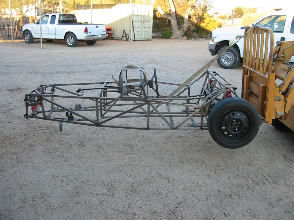Bare steel tube-frame chassis on gravel outdoors with red coilovers and one steel wheel