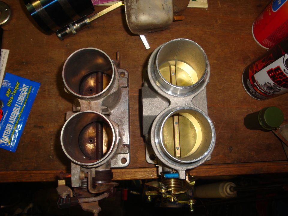 Two throttle bodies on a wooden surface, one appears used and dirty, the other is clean and new