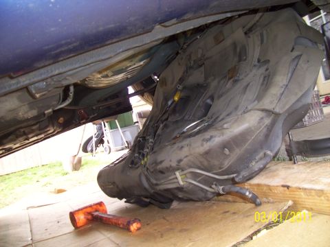 1995 Ford Probe - A vehicle's fuel tank is shown removed from its undercarriage, supported by wooden blocks, with an orange mallet on the ground.