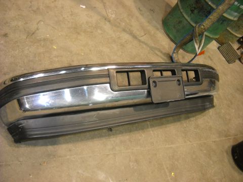 1993 Ford C-150 (Centurion Conversions) - A detached front bumper sits on a concrete floor, with a green barrel and other items in the background.