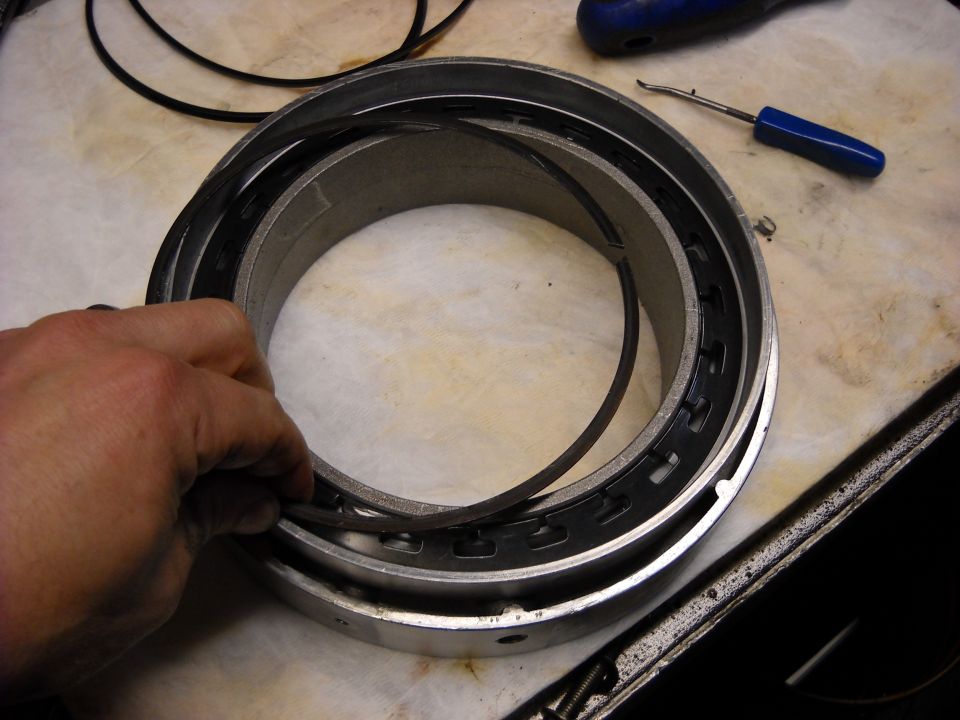 Hand holding a metal ring with black seals inside a larger metal cylinder. Tools are on the table