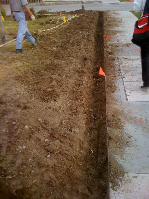 1968 Ford Mustang - Workers dig a trench along a sidewalk, with orange flags marking the area.