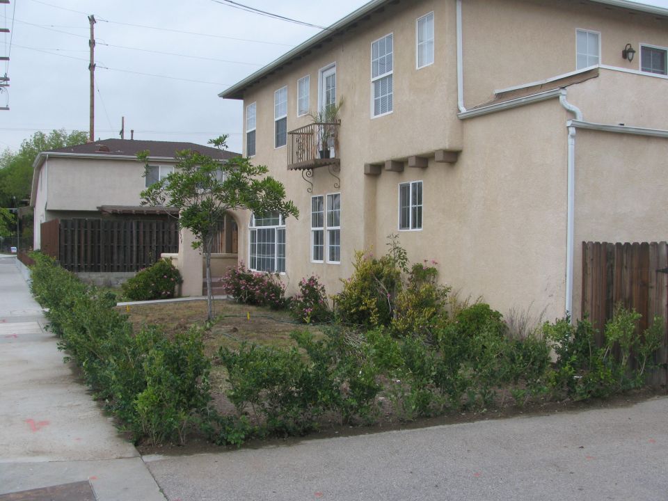 Building with a balcony and a small tree in front, next to a sidewalk and a wooden fence