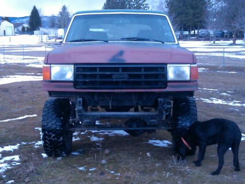 1988 Ford Bronco - A maroon Ford Bronco with a black grille and oversized tires is parked outdoors in a grassy area with scattered snow.