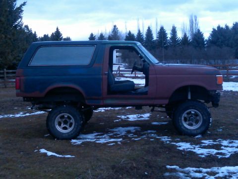 1988 Ford Bronco - This lifted Ford Bronco has its doors removed and sits in a field with scattered snow under a cloudy sky.