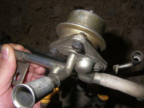 1990 Ford Bronco - A hand holds the end of a steel fuel rail crossover tube with the round canister-style fuel pressure regulator bolted to it, its diaphragm…