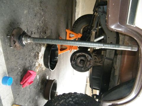 1982 Ford Bronco - A rear axle assembly and brake drum are shown removed from a brown SUV, with tools and parts scattered on the garage floor.