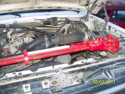 1989 Ford Bronco - The engine bay of a white Ford truck is shown with a red support bar. Another vehicle is visible in the background.
