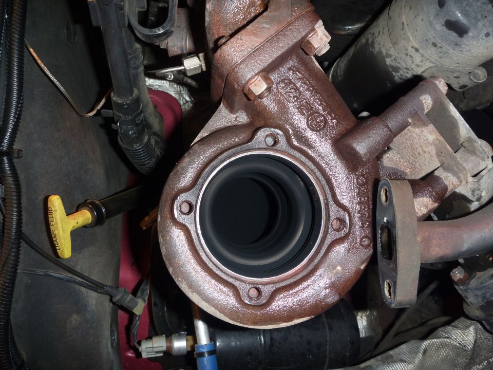 Rusty turbocharger exhaust housing with downpipe removed, yellow trans fluid dipstick visible at left