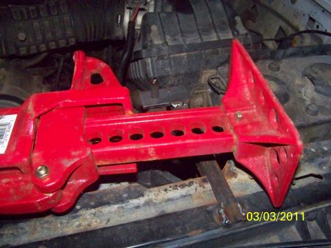 1989 Ford Bronco - Close-up of a red high-lift farm jack resting on a rusty frame rail inside an engine bay, its perforated I-beam bar and cast base plate cle…