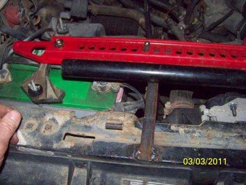 1989 Ford Bronco - Close-up of a red high-lift jack bar and its black steel sleeve laid across the front core support of a truck, with a welded upright tab be…