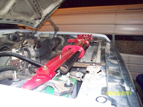 1989 Ford Bronco - Red high-lift jack positioned lengthwise along the front core support of a white truck at night with the hood raised, its base plate and a…