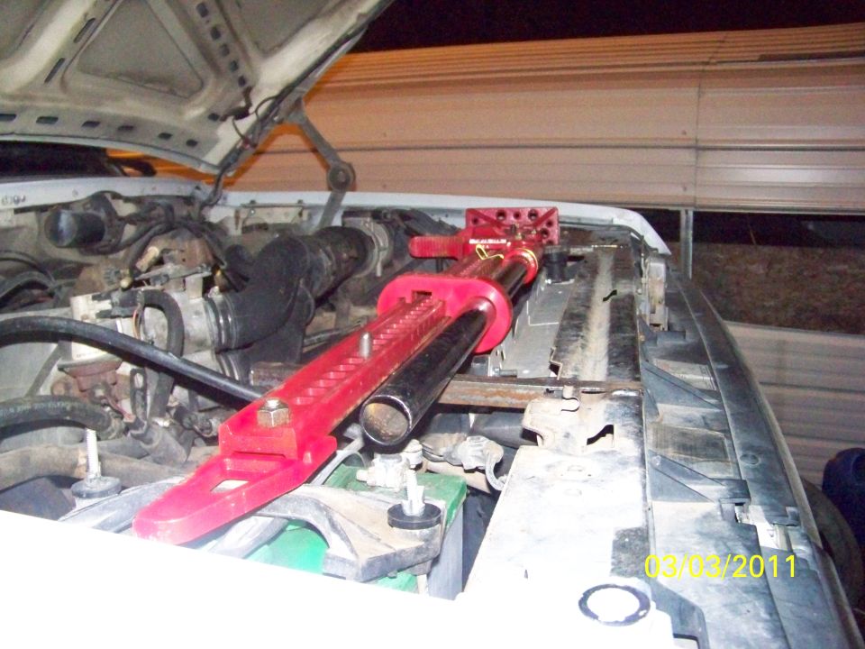 Red high-lift jack mounted along the core support of a white truck with hood up