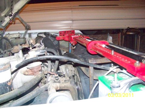 1989 Ford Bronco - A red high-lift farm jack laid diagonally across an open engine bay, its foot resting on a bolt-hole mounting plate at the front of the tru…
