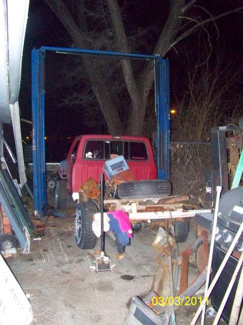 1989 Ford Bronco - Night view of a cluttered outdoor work yard with a blue two-post lift frame straddling a red Ford pickup cab. In the foreground sit a strip…