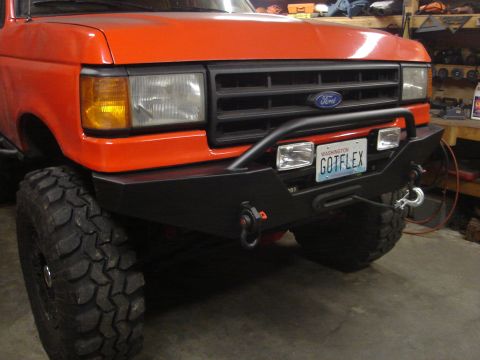 1989 Ford Bronco - Orange Ford truck front with steel winch bumper, mud tires and a Washington GOTFLEX plate in a garage.