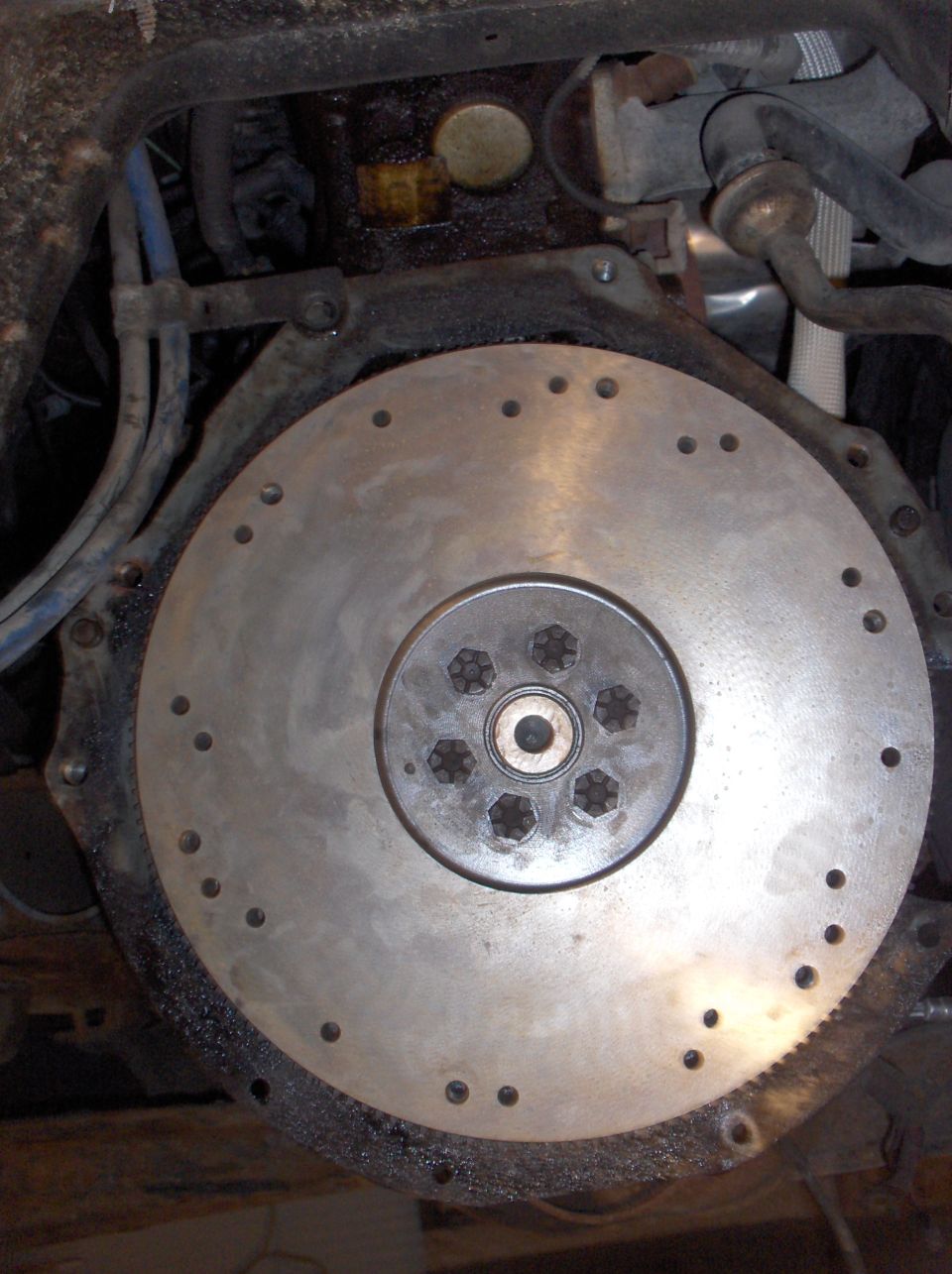 Flywheel with six bolts in the center, surrounded by smaller bolt holes and a toothed edge. Engine components and hoses are visible