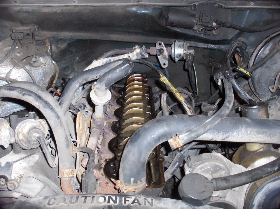 Engine bay showing valve cover, hoses, and wiring. Text 'CAUTION FAN' visible on a part