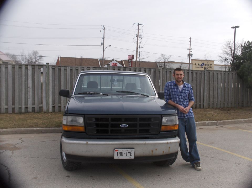 Man leaning on front of dark blue Ford F-150 pickup truck. License plate reads 180-1YE