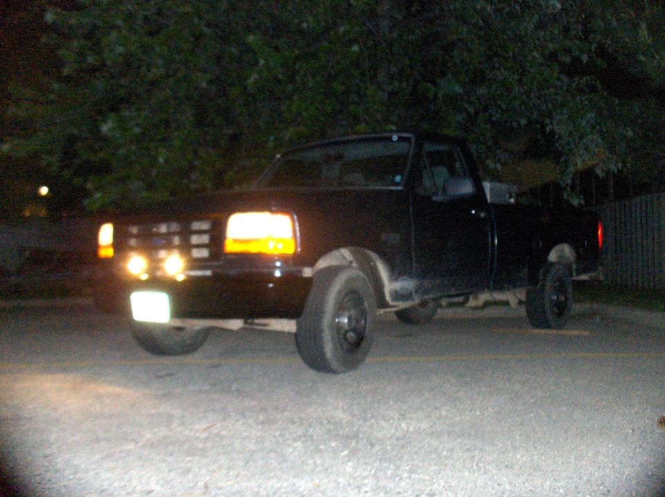Black 1994 Ford F-150 pickup truck parked at night, front end visible with headlights and auxiliary lights on