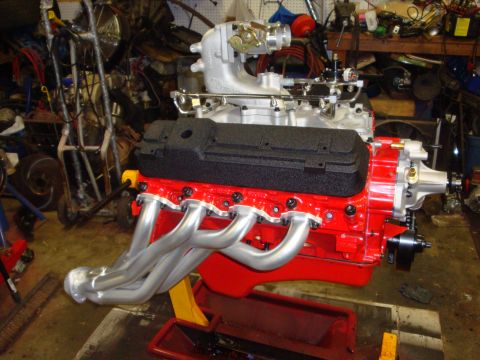 1989 Ford Bronco - A red V8 engine with silver headers and black valve covers sits on a yellow engine stand in a cluttered garage.