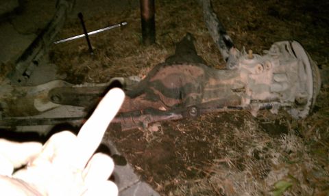 1990 Ford Bronco - A differential axle assembly lies on the ground, partially obscured by grass and dirt. A hand points towards the center of the part.