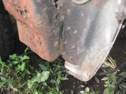 1983 Ford Bronco - Low ground-level view of a rusted rear body corner and rear frame bracket area, with heavy orange scale rust and a split seam where two pan…