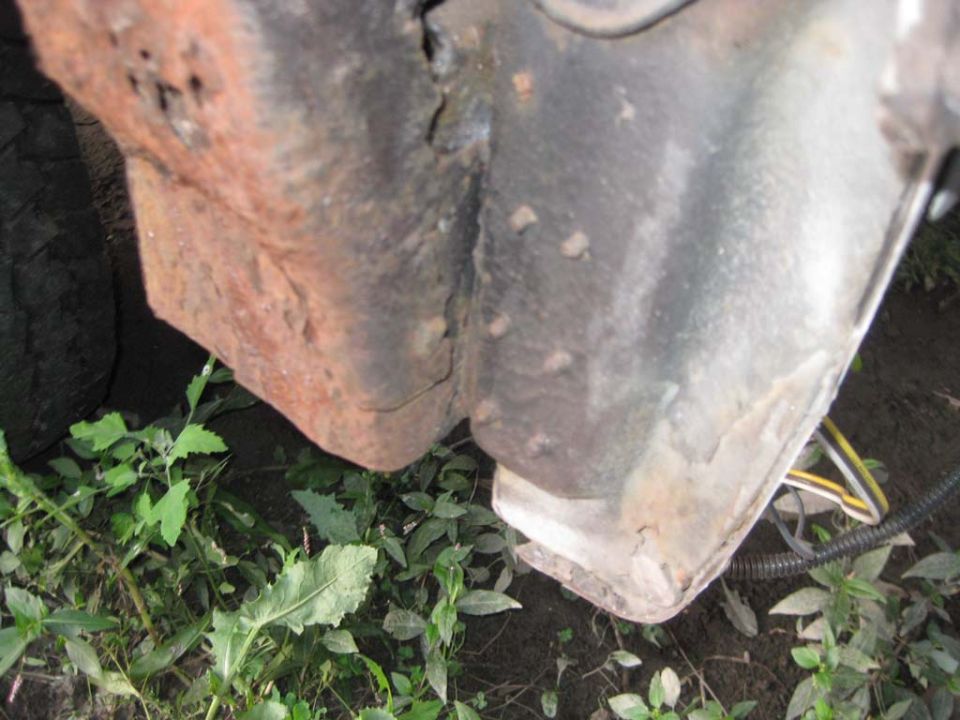 Rusted rear body corner and frame bracket seen from ground level with weeds and wiring