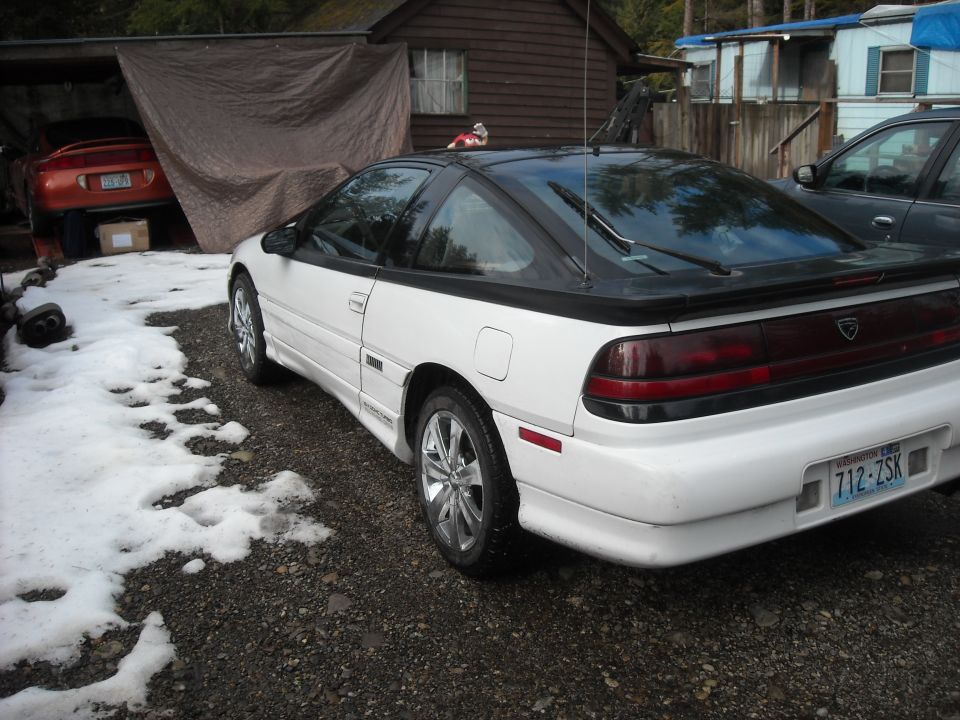 White coupe with black roof and tinted rear glass, rear 3/4 view, parked on snowy gravel.