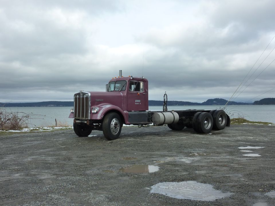 Purple 1959 Kenworth W900A semi-truck chassis parked on gravel by a body of water