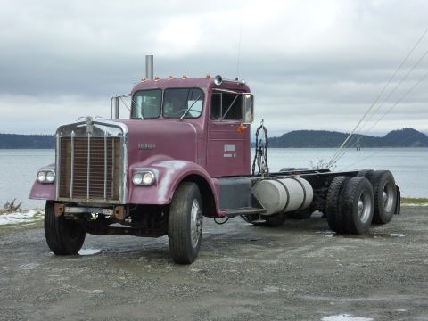 1959 Kenworth W900A - A 1959 Kenworth W900A semi-truck cab and chassis, painted purple, sits parked near a body of water under a cloudy sky.